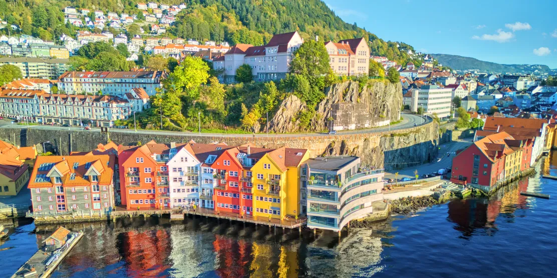 Aerial view of Bergen, Norway, showing colourful wooden waterfront buildings in red, yellow and white built on stilts over the harbour, backed by a forested hillside with clifftop houses.