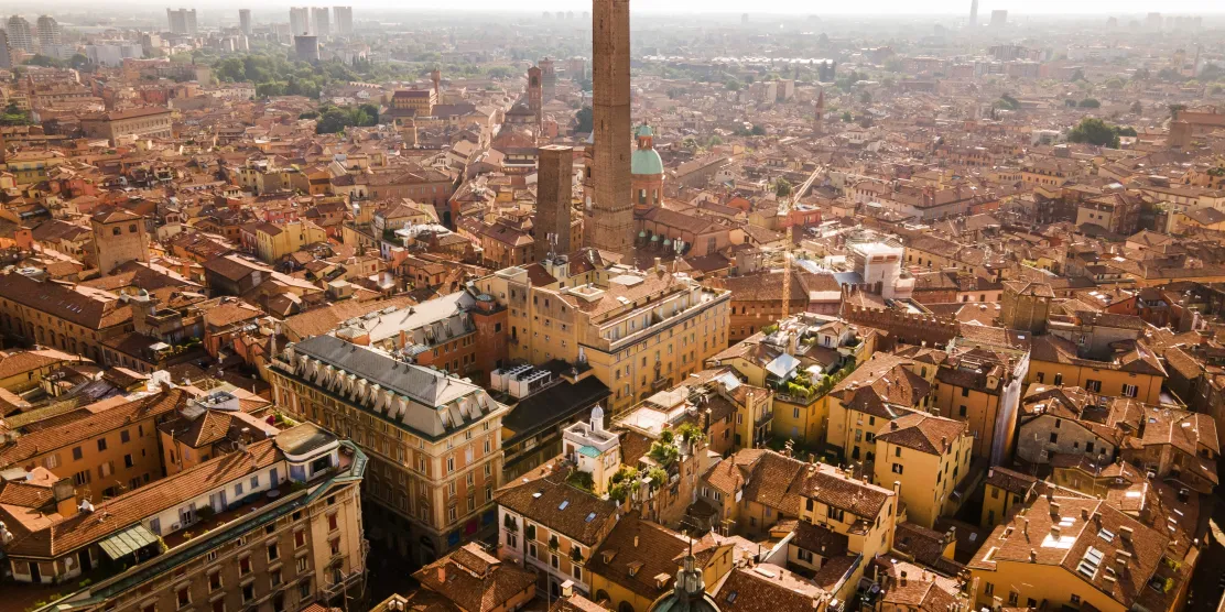 Aerial view of Bologna, Italy, showing the city's iconic leaning medieval towers rising above a dense sea of orange terracotta rooftops.