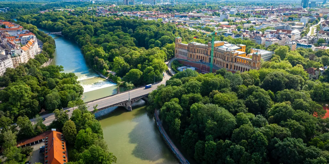 Aerial view of Munich, Germany, showing the Maximilianeum building surrounded by lush green forest along the Isar River, with a bridge crossing the water in the foreground.