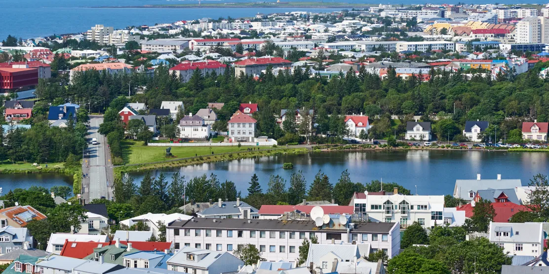 Aerial view of Reykjavik, Iceland, showing a colourful residential neighbourhood with red and blue rooftops surrounding a city pond, with the ocean visible in the background.