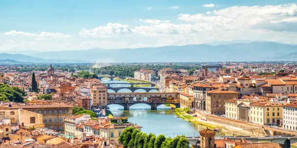 Aerial view of Florence, Italy, showing the Ponte Vecchio bridge spanning the Arno River, surrounded by terracotta rooftops with the Tuscan hills in the background.